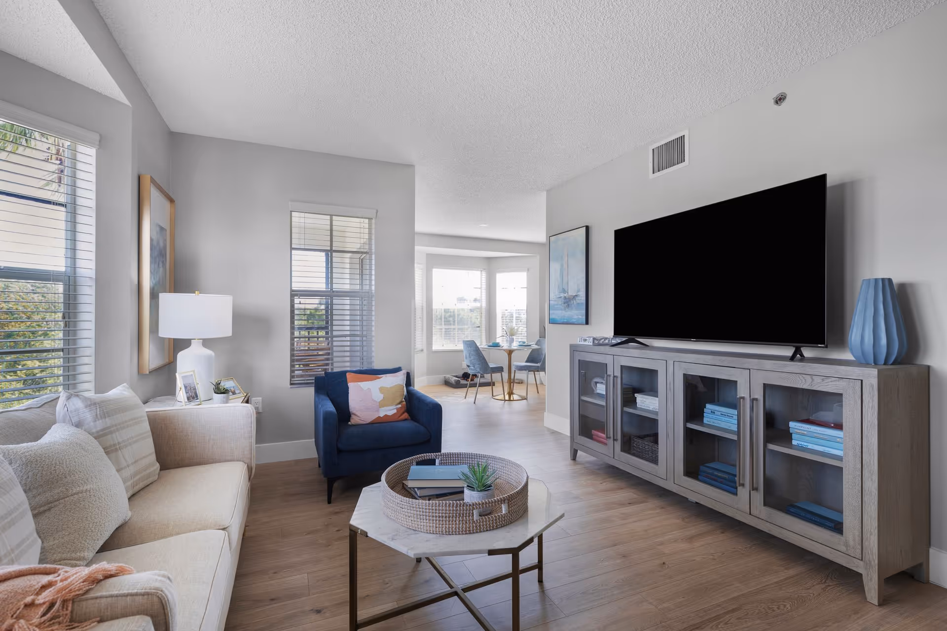 A bright and modern living room with a beige sofa adorned with cushions, a blue armchair with a colorful pillow, a marble-top coffee table with a decorative tray and books, a large flat-screen TV on a wooden cabinet with glass doors, and a dining area with a round table and blue chairs near large windows letting in natural light.