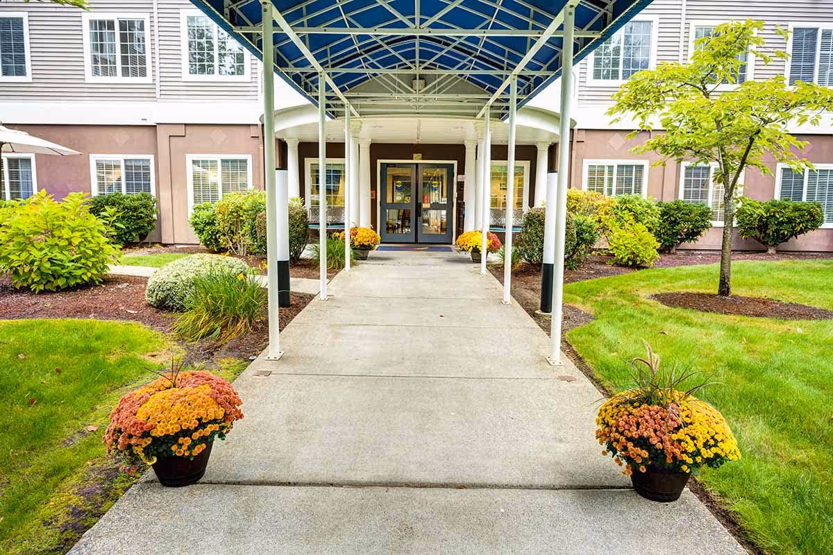 Covered walkway leading to the entrance of a building with glass double doors, surrounded by green shrubs, trees, and flower pots with yellow and orange flowers on either side of the path.