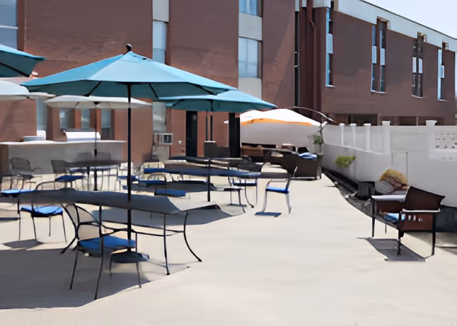 Outdoor patio area with several tables and chairs under large blue and white umbrellas, adjacent to a brick building with multiple windows.