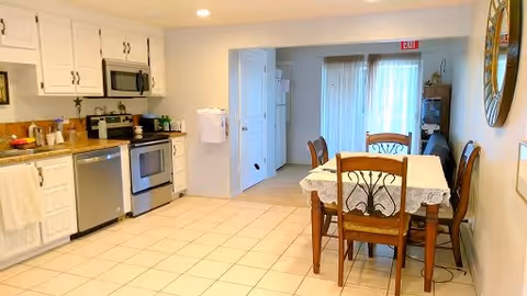 A bright and clean kitchen and dining area in an assisted care home. The kitchen features white cabinets, a dishwasher, an oven with a microwave above it, and various kitchen items on the countertops. The dining area has a wooden table with a lace tablecloth and four chairs. There is a large mirror on the wall and a sliding glass door with sheer curtains leading outside.