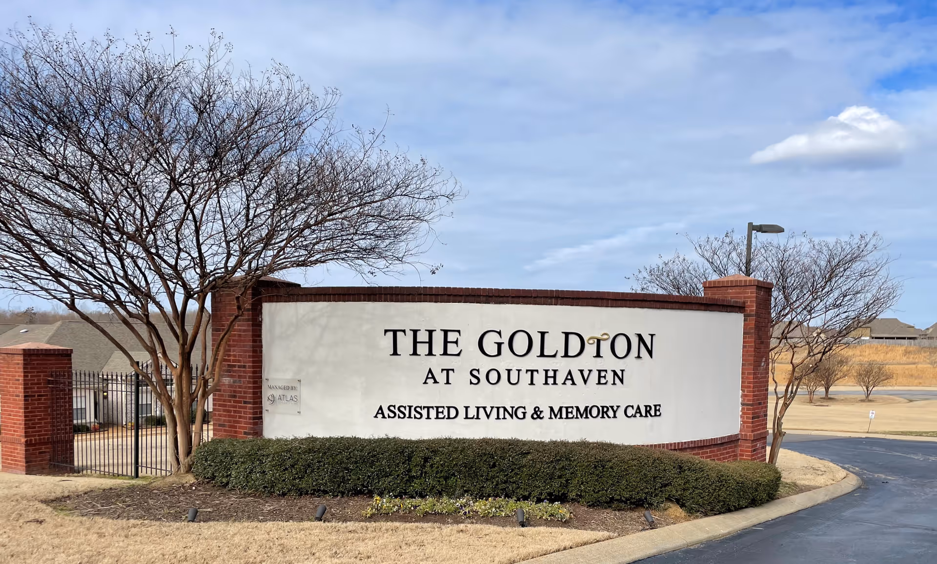 Entrance sign reading "The Goldton at Southaven Assisted Living & Memory Care" set in a brick wall with landscaping, trees, and a driveway.