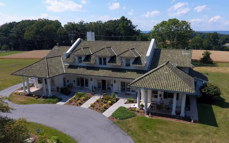 A large single-story building with a green tiled roof and multiple dormer windows, surrounded by landscaped gardens and a curved driveway, set in a rural area with fields and trees in the background under a blue sky with scattered clouds.