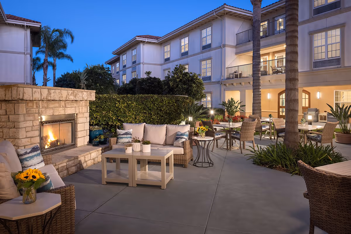 Outdoor patio area at dusk with comfortable wicker seating, a stone fireplace with a fire burning, tables with chairs, potted plants, and a multi-story building in the background with lit windows and balconies.