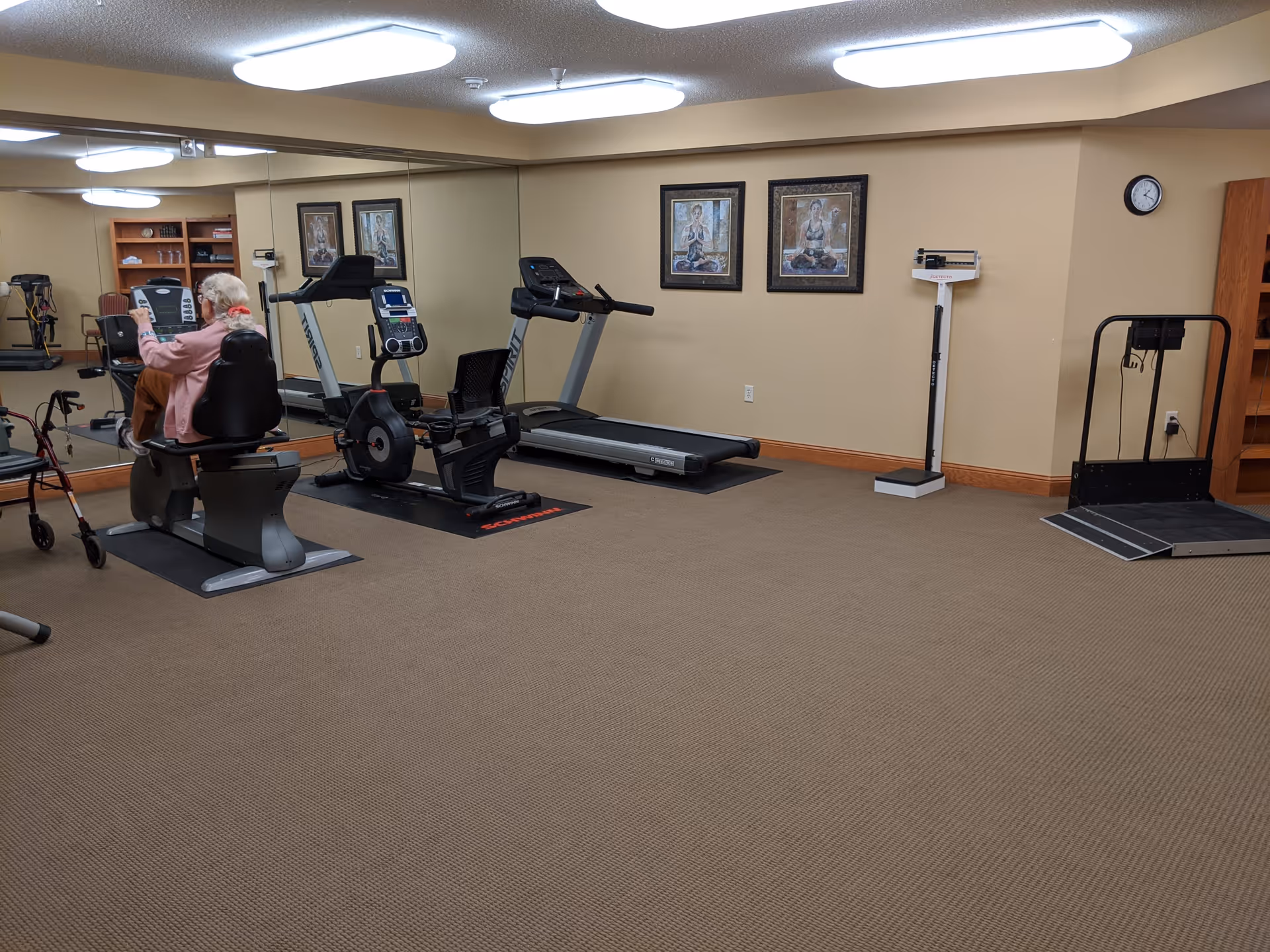 An elderly person using a recumbent exercise bike in a fitness room with a treadmill, rowing machine, weight scale, and a wheelchair ramp. The room has beige walls, carpeted floor, two framed pictures on the wall, a clock, and a large mirror reflecting the equipment.