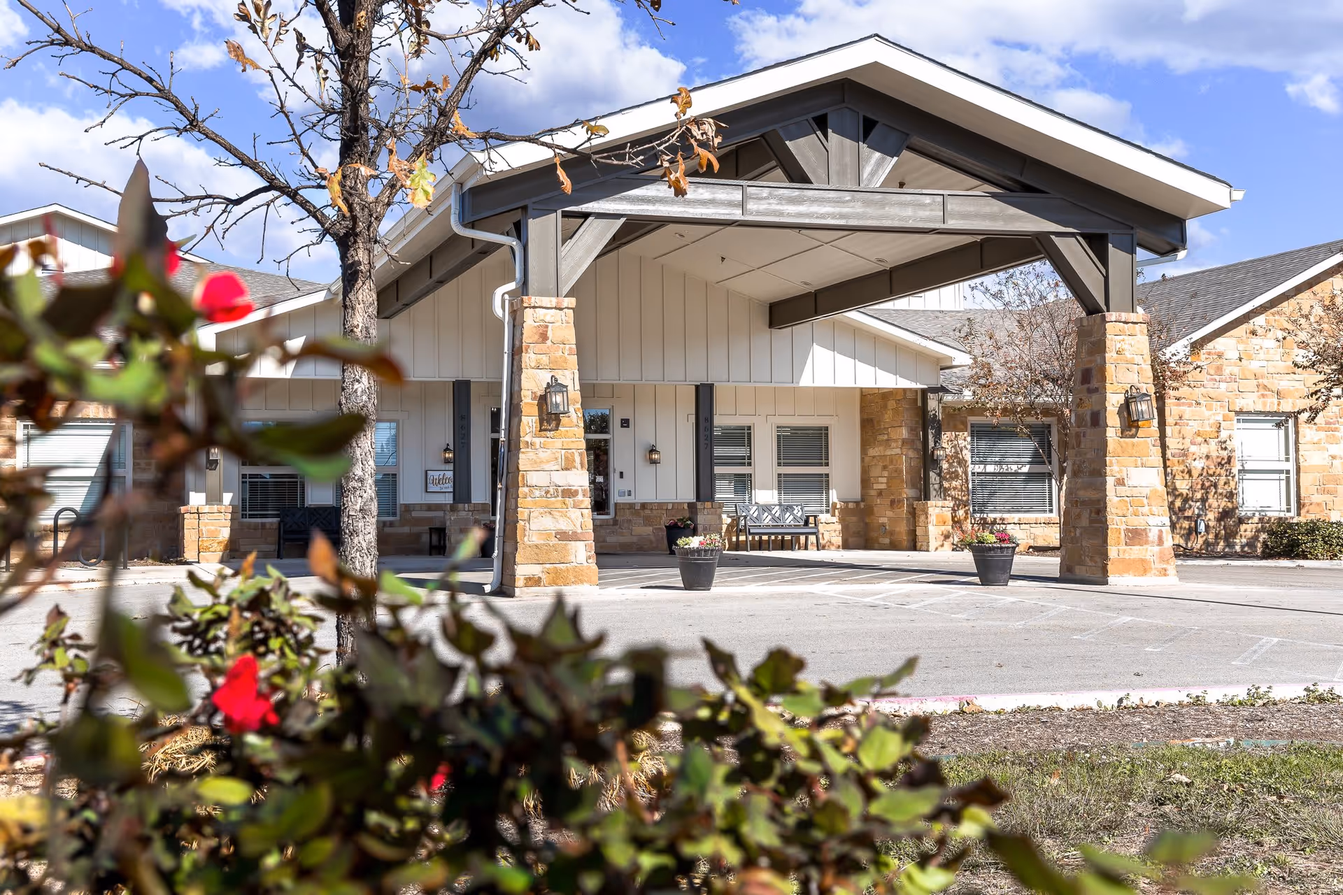 Covered entrance and porte-cochère of a senior living building with stone pillars, benches, and landscaped foreground.