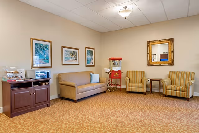 A cozy lounge with a sofa, two striped armchairs, a popcorn machine, a snack cabinet, framed artwork, and a wall mirror.
