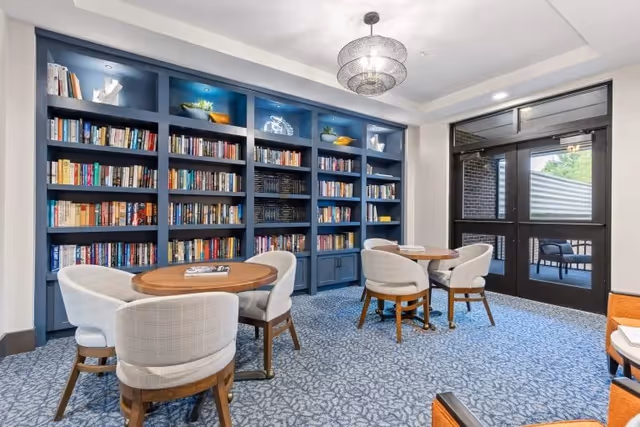 A cozy reading room with blue built-in bookshelves filled with books and decorative items. There are two round wooden tables, each surrounded by four cushioned chairs with wooden legs. The room has a patterned blue carpet and a modern chandelier hanging from the ceiling. A glass door leads to an outdoor area with additional seating visible.
