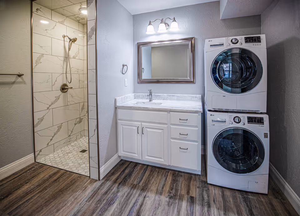A modern bathroom featuring a walk-in shower with marble-patterned tiles and a handheld showerhead, a white vanity with a marble countertop and a rectangular mirror above it, and a stacked washer and dryer unit to the right. The floor has wood-like vinyl planks and the walls are painted light gray.