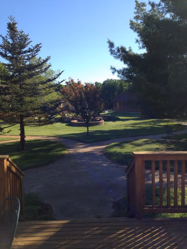View from a wooden deck overlooking a well-maintained outdoor area with green grass, trees, a circular raised flower bed, and a pathway leading to a building in the background under a clear blue sky.