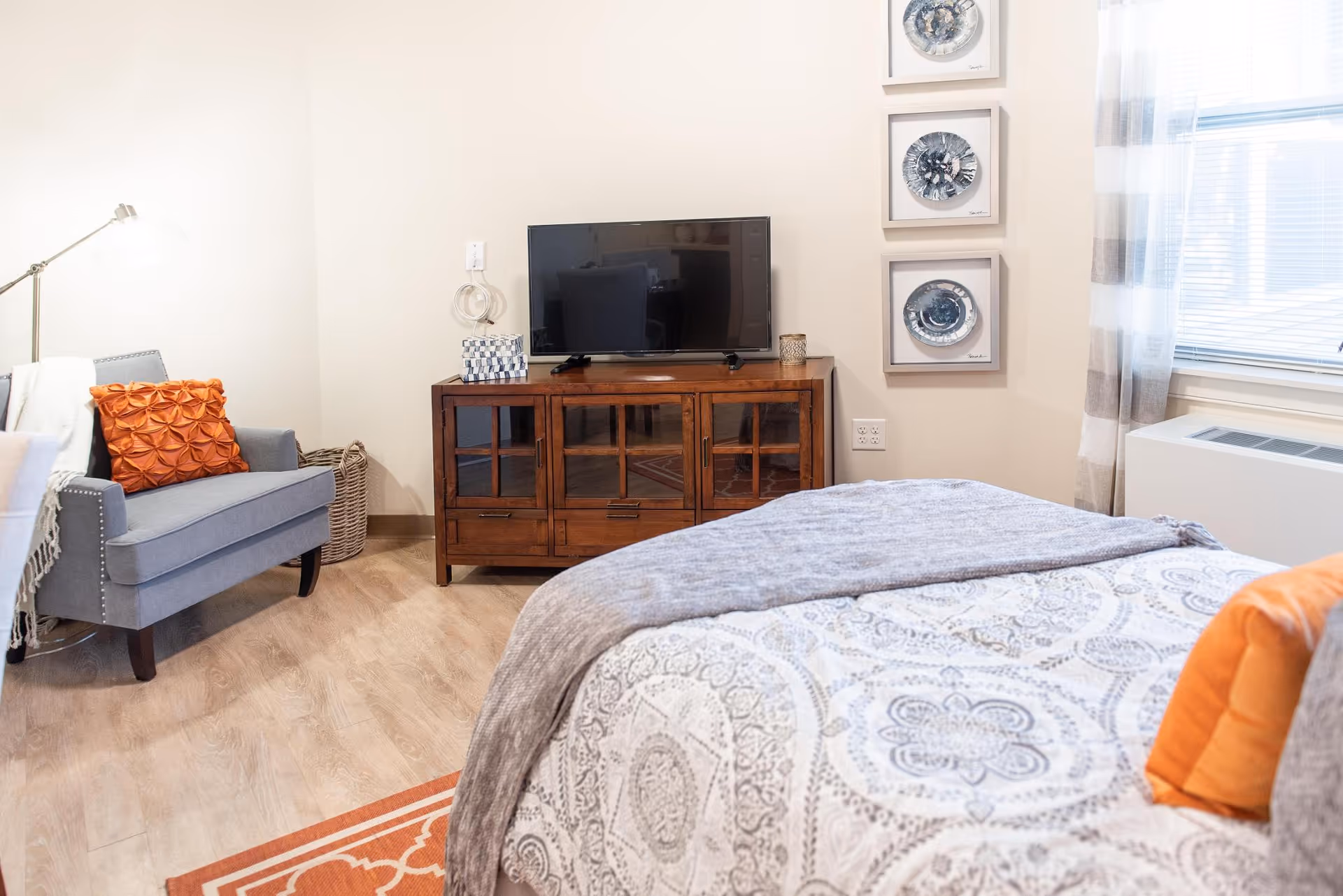 Well-lit furnished bedroom with a bed in the foreground, an armchair, a wooden TV cabinet with a flat-screen, and decorative wall art.