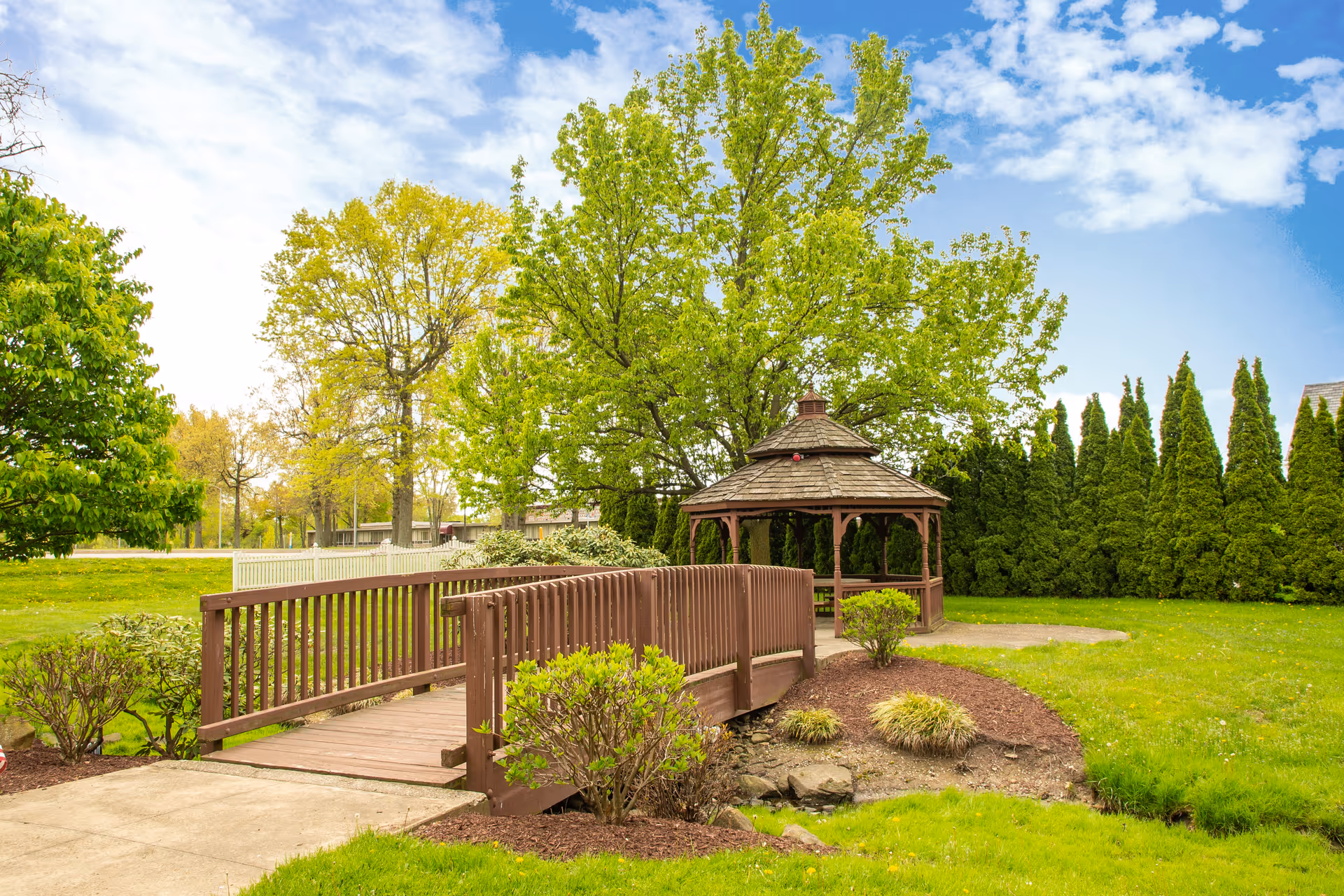 A peaceful outdoor garden area featuring a wooden bridge over a small landscaped section, leading to a wooden gazebo surrounded by green grass, bushes, and tall trees under a partly cloudy blue sky.