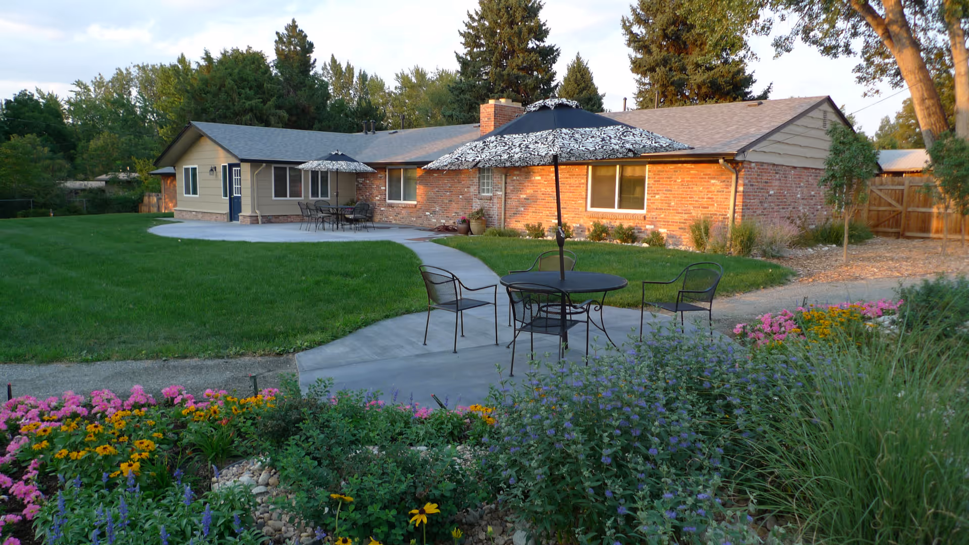Outdoor patio area at a senior living facility with a brick building in the background. The patio has round tables with umbrellas and metal chairs. There is a well-maintained lawn and colorful flower beds surrounding the patio.