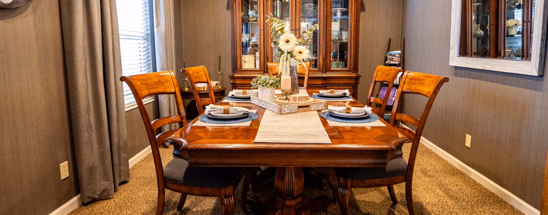 A dining room with a wooden dining table set for four people. The table has a beige runner, blue placemats, white plates with napkins and napkin rings, and a centerpiece with flowers and candles. There are four wooden chairs with cushioned seats around the table. In the background, there is a wooden china cabinet with glass doors displaying decorative items. The room has beige carpet, grayish walls, a window with closed blinds and curtains, and a large mirror on the right wall.