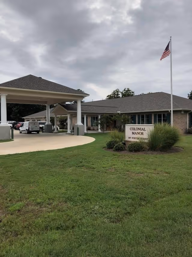 Front entrance of Colonial Manor Nursing & Rehab with a covered drive-up, lawn sign, flagpole, and parked vehicles under a cloudy sky.
