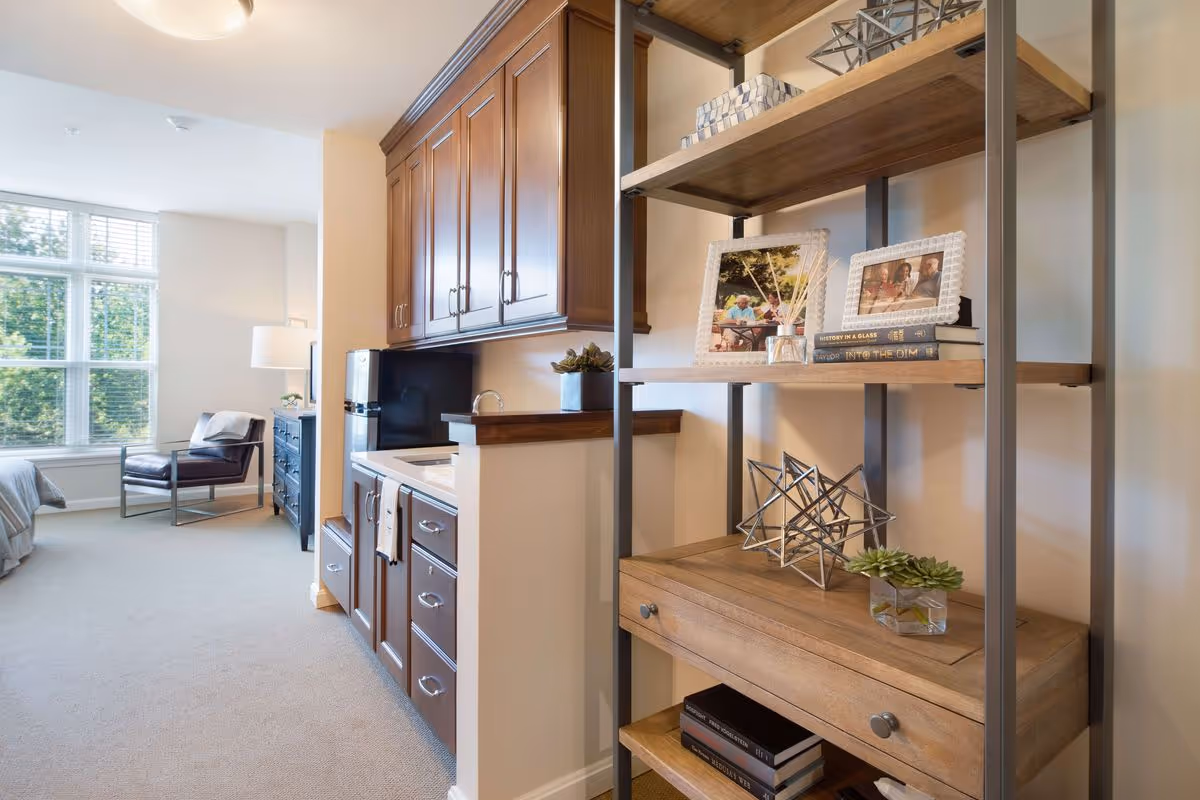 Interior view of a senior living facility room featuring a kitchenette with wooden cabinets and a small sink. To the right, there is a wooden shelving unit with decorative items including framed photos, books, and small plants. In the background, there is a seating area with a chair and a large window letting in natural light.