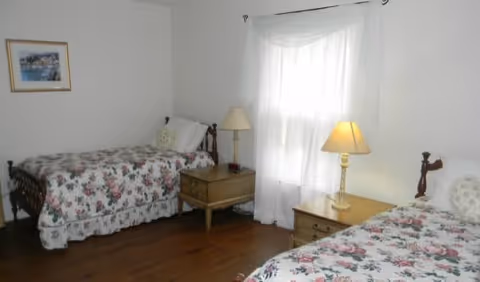 Bright bedroom with two twin beds covered in floral bedspreads, wooden nightstands with lamps, and a window with sheer curtains.