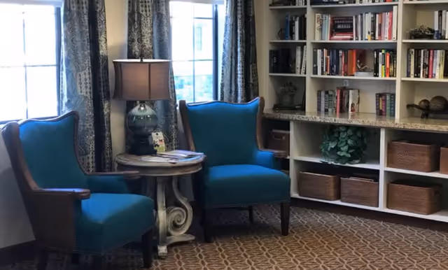 A cozy interior corner with two blue upholstered armchairs flanking a small round wooden table with a lamp and some magazines. Behind the chairs is a window with patterned curtains. To the right, there is a built-in white bookshelf filled with books, decorative items, and woven baskets on the lower shelves. The floor is carpeted with a patterned design.