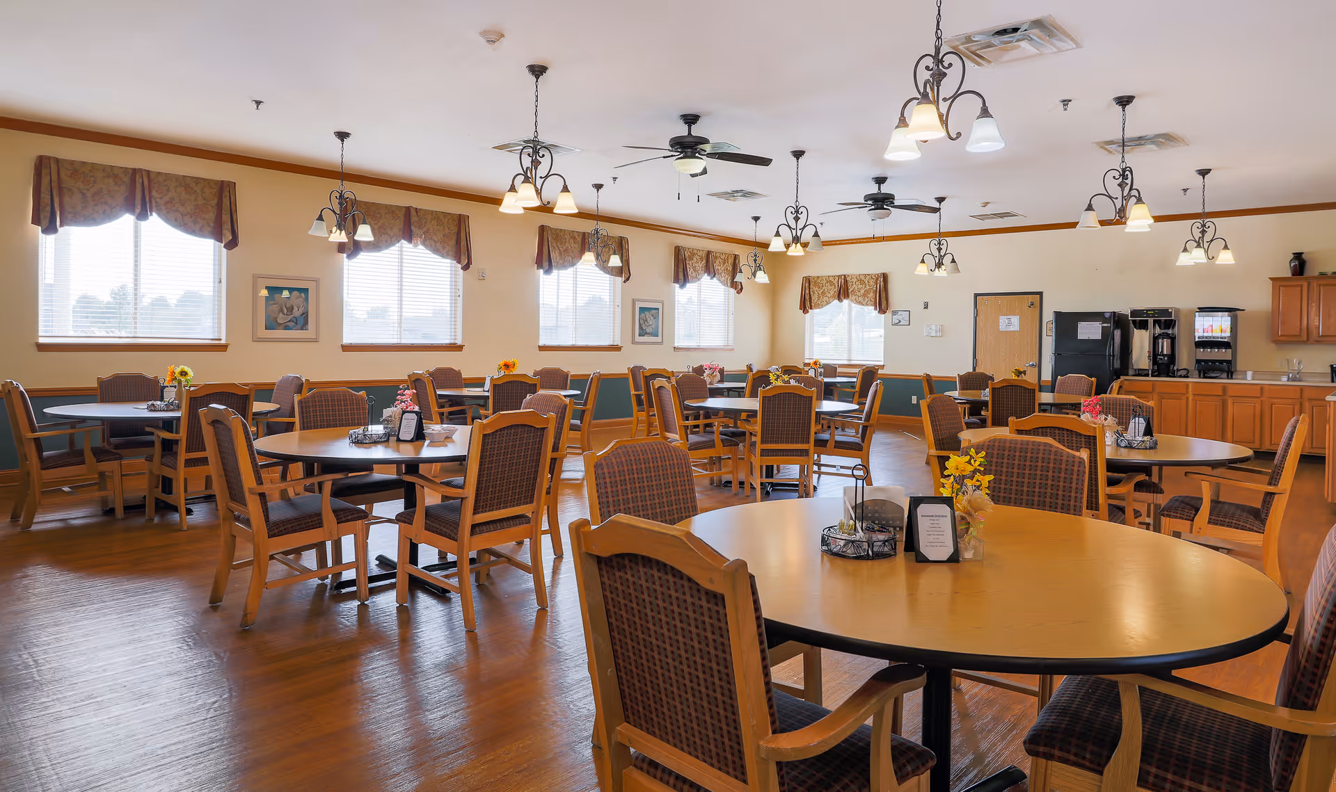 A spacious dining room with multiple round tables and wooden chairs arranged neatly. The room has large windows with valance curtains allowing natural light to fill the space. Ceiling fans and hanging light fixtures provide additional lighting. There is a counter area with coffee machines and cabinets along one wall.