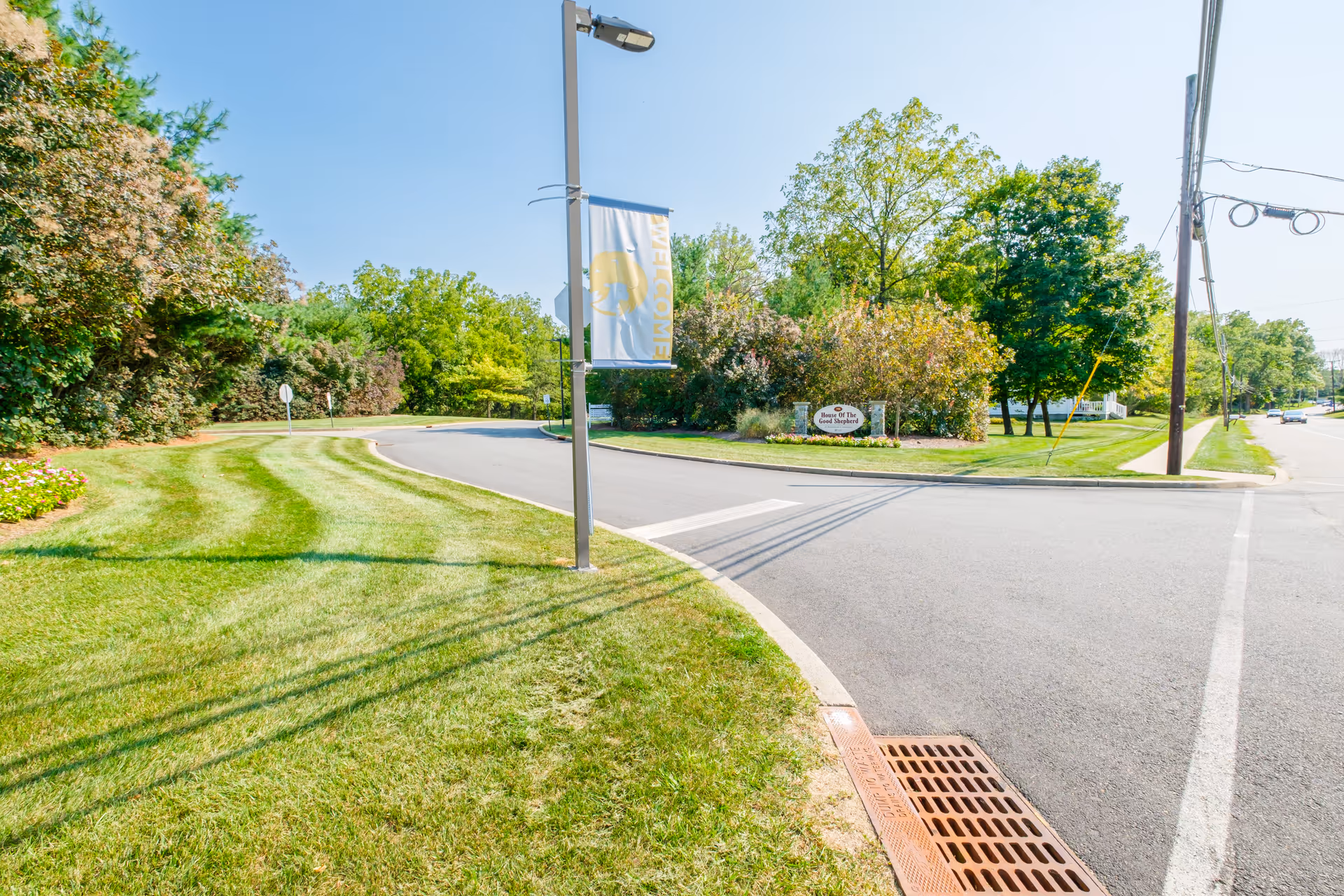 Curving driveway entrance with manicured lawn, trees, and an entrance sign for the senior living community.