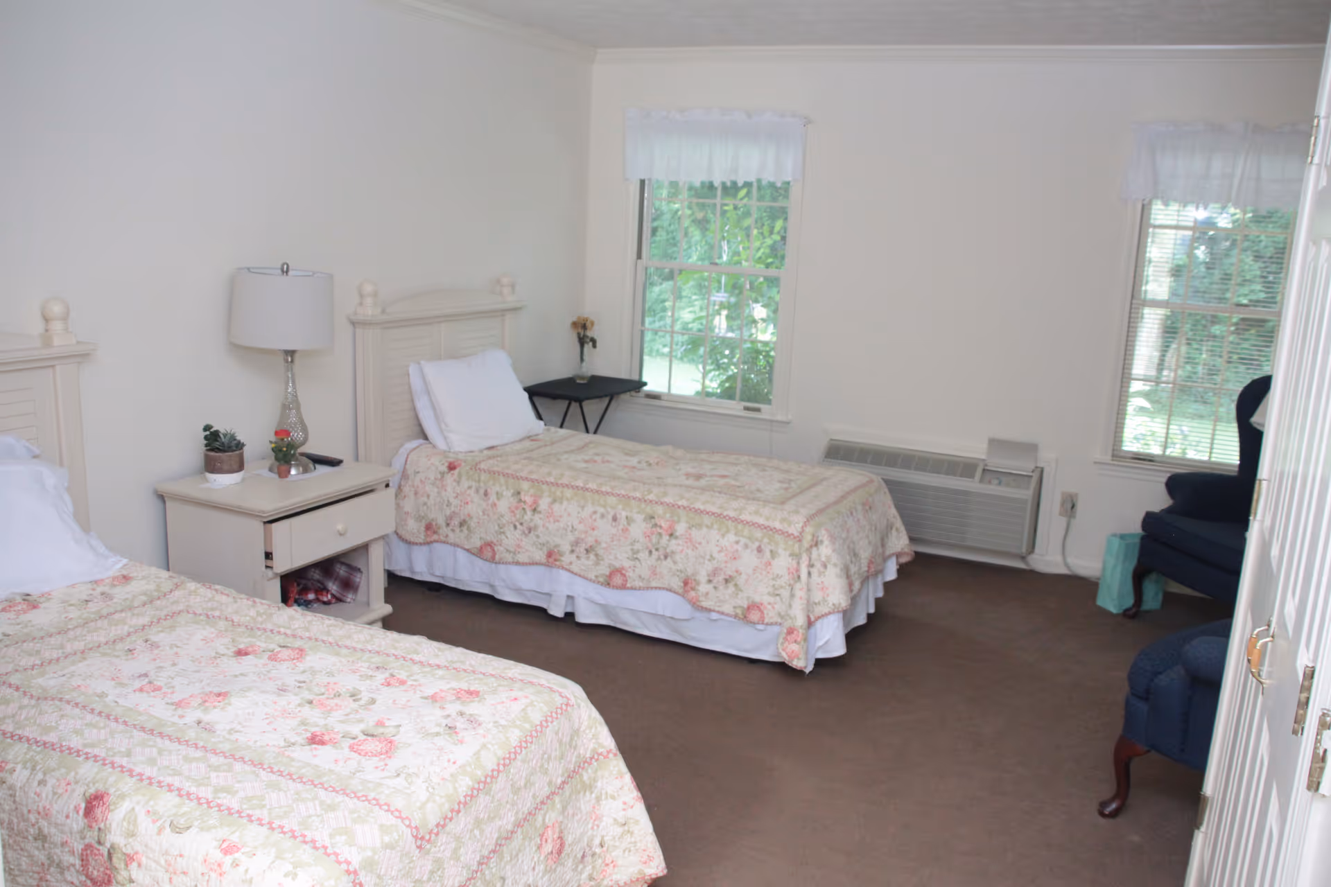 Two twin beds with floral quilts in a softly lit bedroom with a nightstand, lamp, windows, and armchairs.