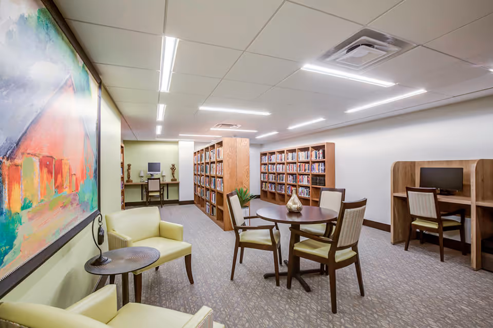 A well-lit library or reading room with bookshelves filled with books, a round table with four chairs, two armchairs with a small round side table between them, a colorful abstract painting on the wall, and computer workstations with chairs along the walls.