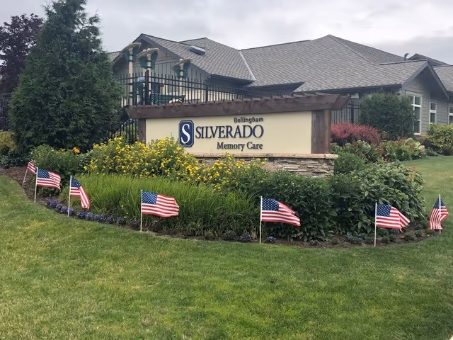 Entrance sign for Silverado Bellingham Memory Care with landscaping, American flags, and the facility building behind it.