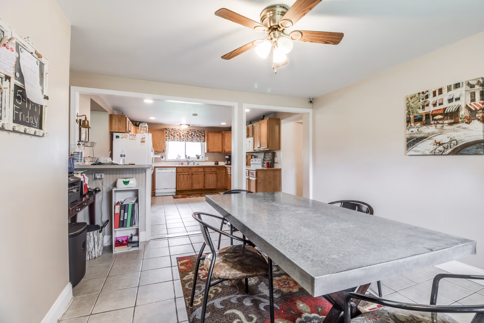 Interior view of a dining area with a rectangular table and four chairs on a patterned rug, adjacent to a kitchen with wooden cabinets, a white refrigerator, dishwasher, and a window with floral curtains. A ceiling fan with lights is on the ceiling, and a painting of a street scene hangs on the wall.