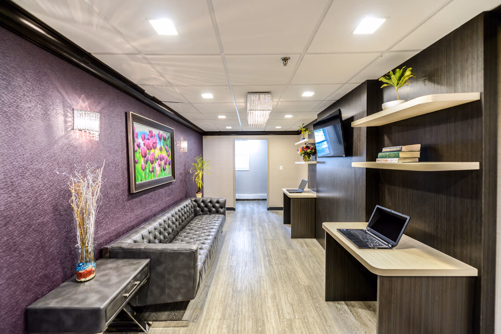 A modern interior lounge area with a long black leather tufted sofa along a purple textured wall. Above the sofa is a framed picture of pink flowers. On the opposite wall, there are two wooden desks with laptops, floating shelves with books and plants, and a mounted flat-screen TV. The floor is light wood, and the ceiling has recessed lighting and a small chandelier.