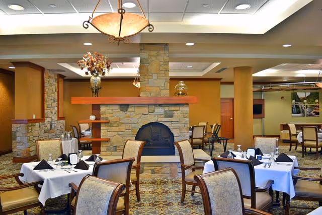 A dining room with several tables covered in white tablecloths, each set with black napkins, plates, and silverware. The room features a stone fireplace in the center with a wooden mantel, elegant chandeliers hanging from the ceiling, and upholstered chairs around the tables. The floor has a patterned carpet, and the walls are painted in warm tones.