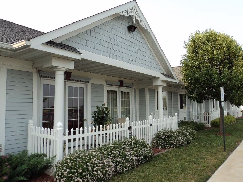 Exterior view of a single-story building with light blue siding and white trim, featuring a covered porch with white columns and a white picket fence. There are flowering bushes and a tree along a sidewalk in front of the building.