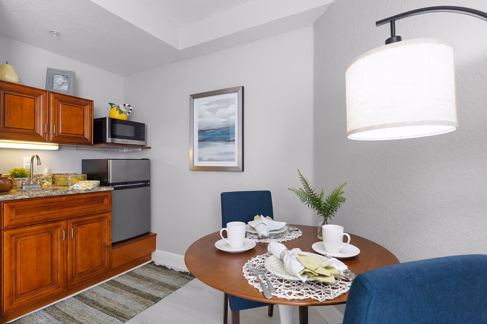 Small kitchenette with wooden cabinets, a mini fridge and microwave beside a round dining table set for two under a hanging lamp.