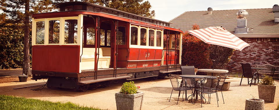 Outdoor patio area with metal tables and chairs, one table shaded by a red and white striped umbrella, next to a vintage red and cream trolley car labeled 'Knott's Berry Farm'. The scene includes potted plants and a brick building in the background.
