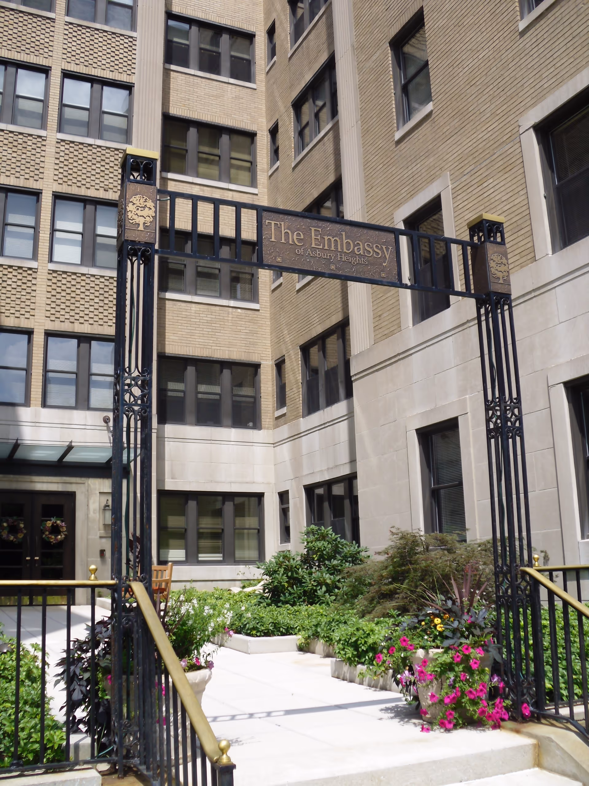 Entrance courtyard with a decorative iron gate reading "The Embassy of Asbury Heights" in front of a multi-story brick building and landscaped planters.