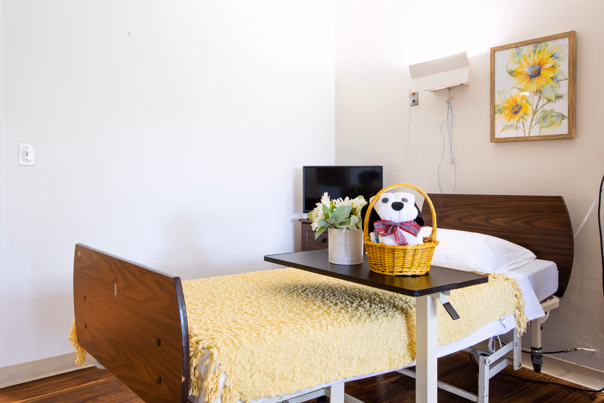 A neatly made hospital-style bed with a yellow blanket and a white pillow in a room. On the bed is a small table holding a basket with a stuffed animal and a vase with flowers. A small TV is on a stand against the wall, and a framed painting of sunflowers hangs above the bed.