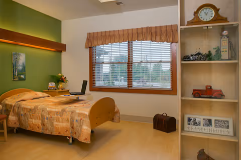 A cozy bedroom in a senior living facility with a single bed covered in a patterned quilt, a small bedside table with a lamp and flowers, a laptop on the bed, a large window with blinds and a valance, and a shelving unit with decorative items including a clock, toy truck, and framed photos.