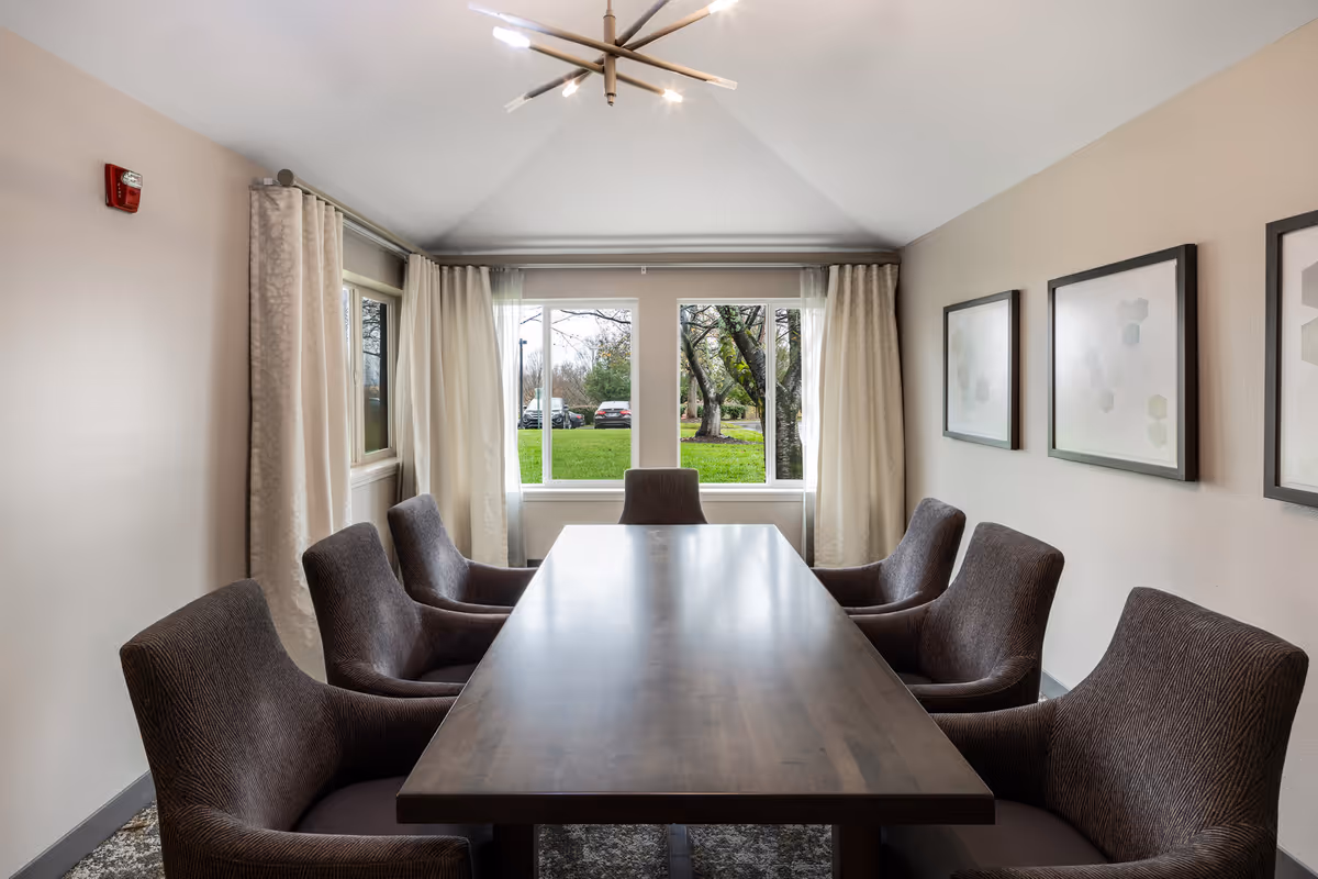 A small conference or dining room with a long wooden table surrounded by eight upholstered chairs. The room has beige walls with three framed abstract art pieces on one side and large windows with cream-colored curtains letting in natural light. Outside the windows, green grass and trees are visible.