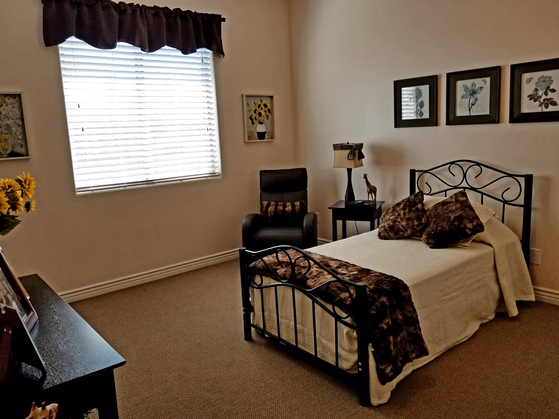 A cozy bedroom with a single bed featuring a black metal frame, white bedding, and brown fur-like pillows and throw. There is a black armchair with a matching pillow next to a small black side table holding a lamp and a small giraffe figurine. The walls are decorated with framed floral artwork, and a window with white blinds and a dark valance lets in natural light. A black dresser with picture frames and a vase of sunflowers is partially visible.