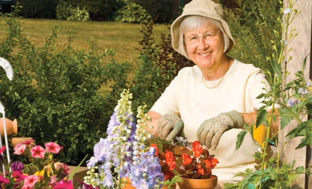 An elderly woman wearing a beige hat and gloves is gardening outdoors, smiling while tending to red flowers in a pot surrounded by various colorful flowers and green plants.