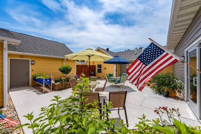 Sunny outdoor courtyard with patio tables, umbrellas, potted plants, and an American flag between yellow buildings.