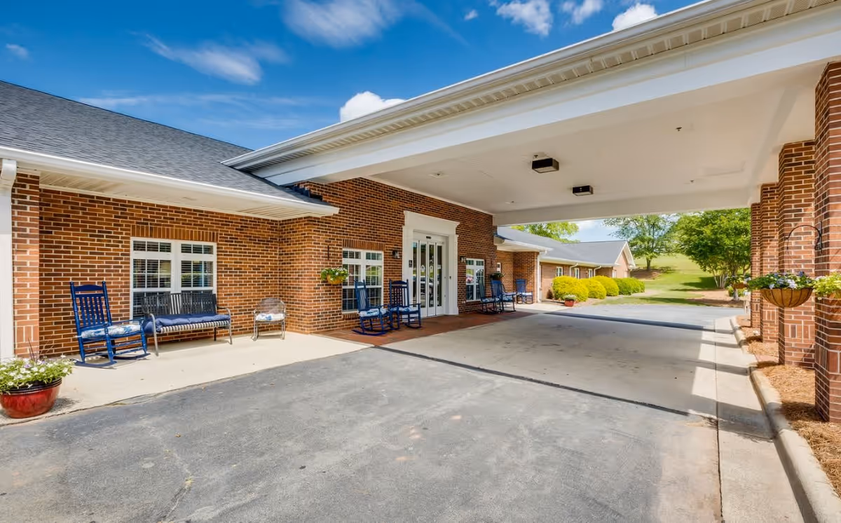 Entrance area of Walnut Ridge Assisted Living featuring a covered drop-off zone with brick walls, several blue rocking chairs and benches along the wall, potted plants, and a clear blue sky with some clouds.