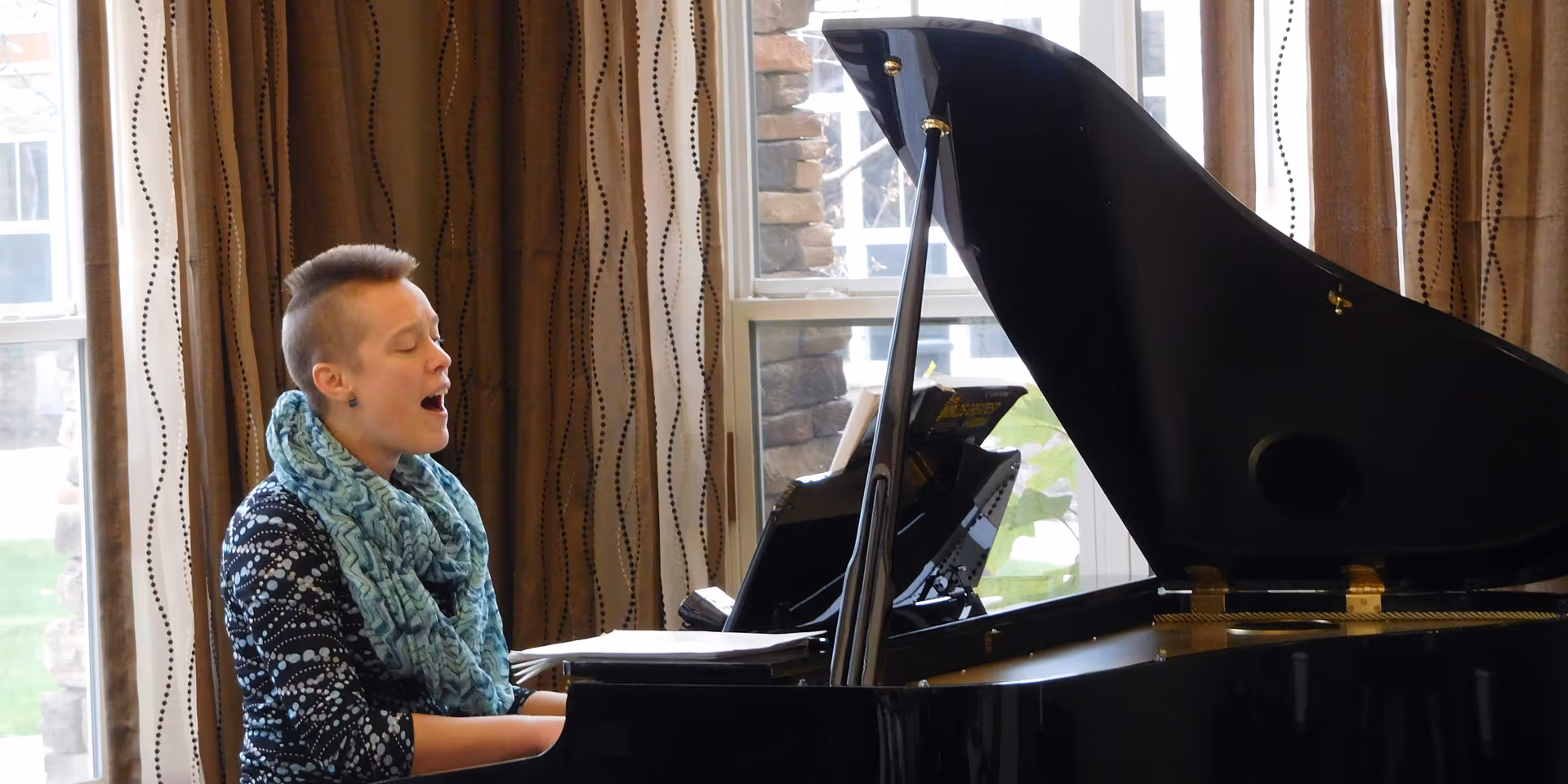 A person with short hair and a blue patterned scarf is playing a grand piano and singing in a room with large windows and brown curtains.