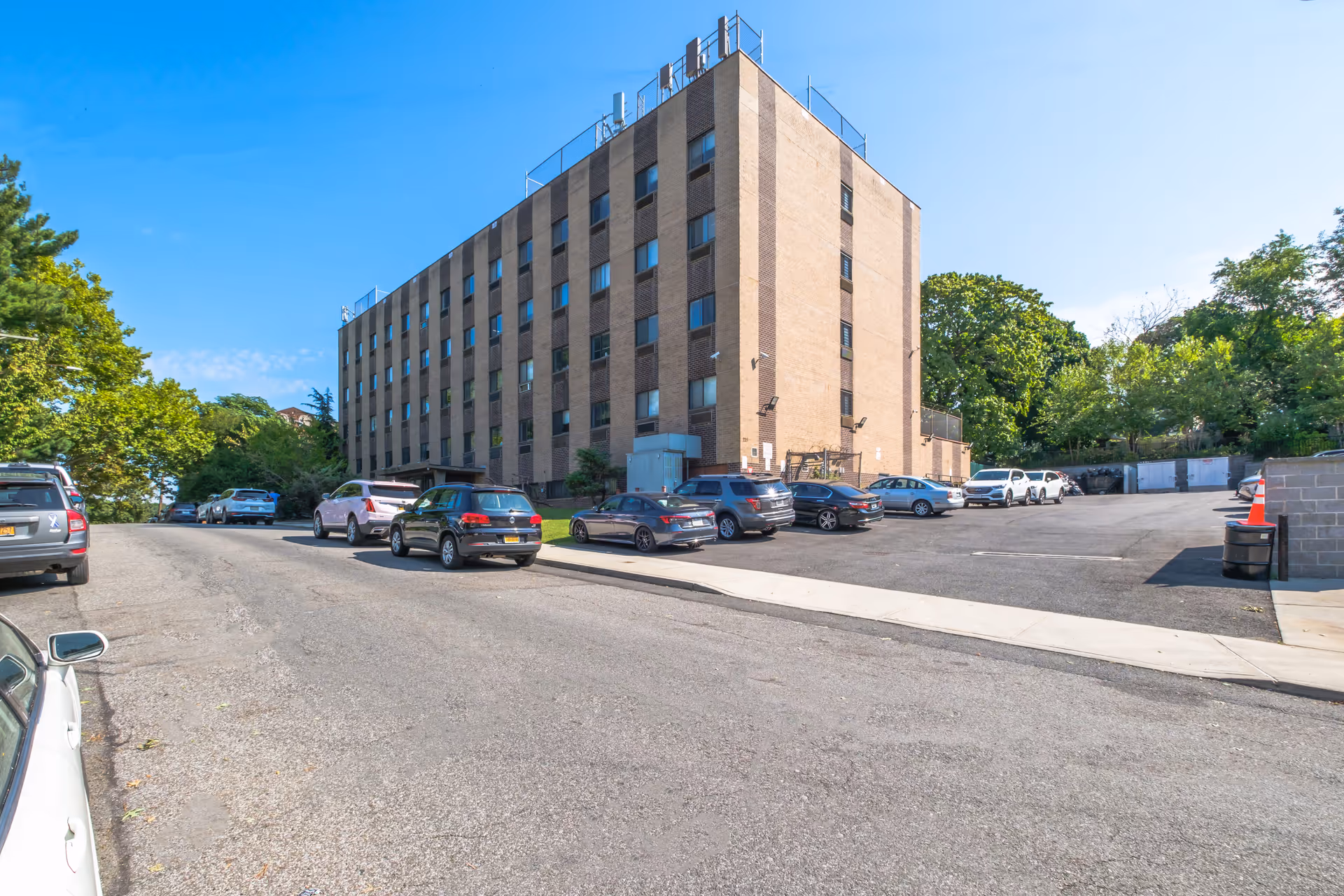 A multi-story brick building with a paved parking lot and cars parked in front, surrounded by trees.