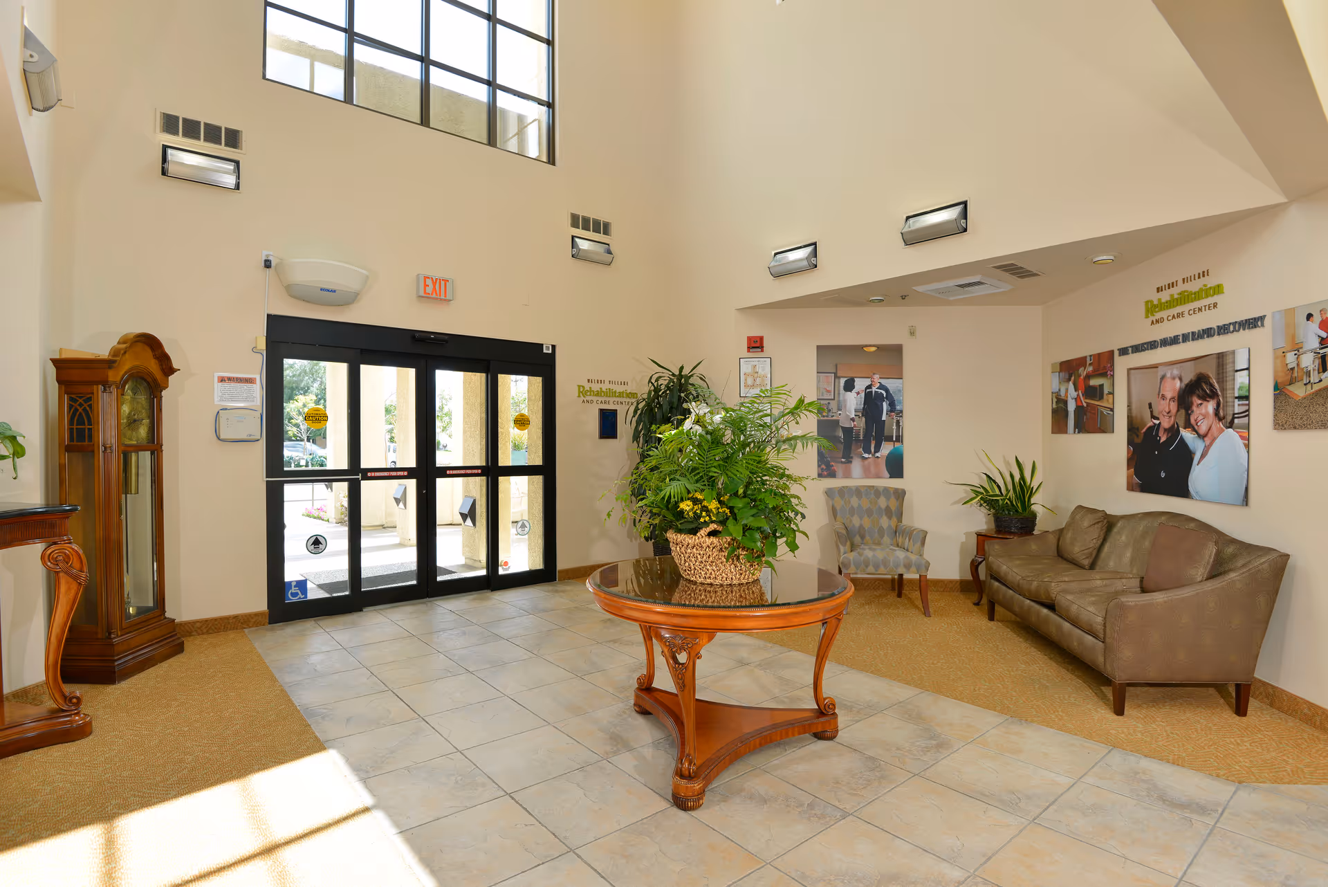 Bright and spacious lobby area of Walnut Village Orange County Retirement Community featuring a large glass entrance door, a round wooden table with a plant centerpiece, a grandfather clock, a sofa, an armchair, and several framed photos and informational posters on the walls.
