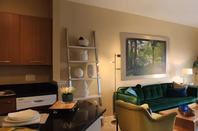 A cozy living room area with a green velvet sofa, a yellow armchair, a wooden coffee table with decorative items, and a large framed picture of a forest scene on the wall. To the left, part of a kitchen area is visible with wooden cabinets, a dishwasher, and a black countertop with plates and a small plant. A modern ladder-style shelf with white decorative vases stands between the kitchen and living room.