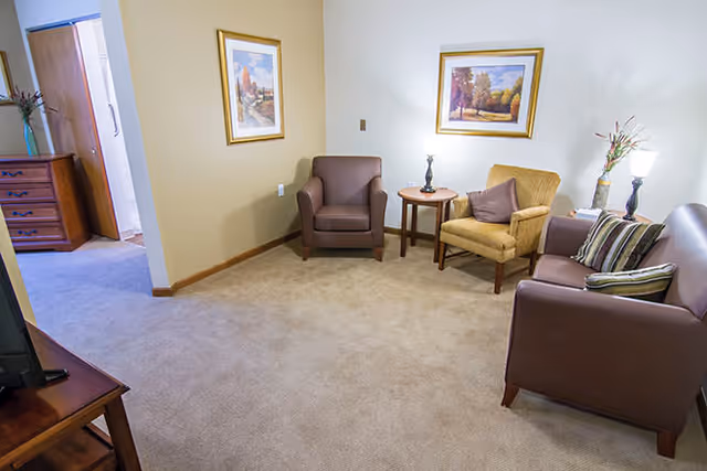 A small living room area with beige carpet and light-colored walls. The room contains a brown leather armchair, a yellow upholstered armchair with a pillow, a brown leather sofa with striped pillows, and a small wooden side table with a lamp. Two framed landscape paintings hang on the walls. A wooden dresser is visible in an adjacent room through an open doorway.