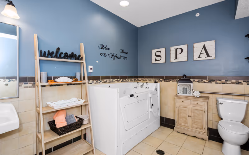 Spa-style bathroom with a walk-in tub, toilet, sink, shelving with towels and decor, and wall signs reading 'SPA' and 'welcome'.