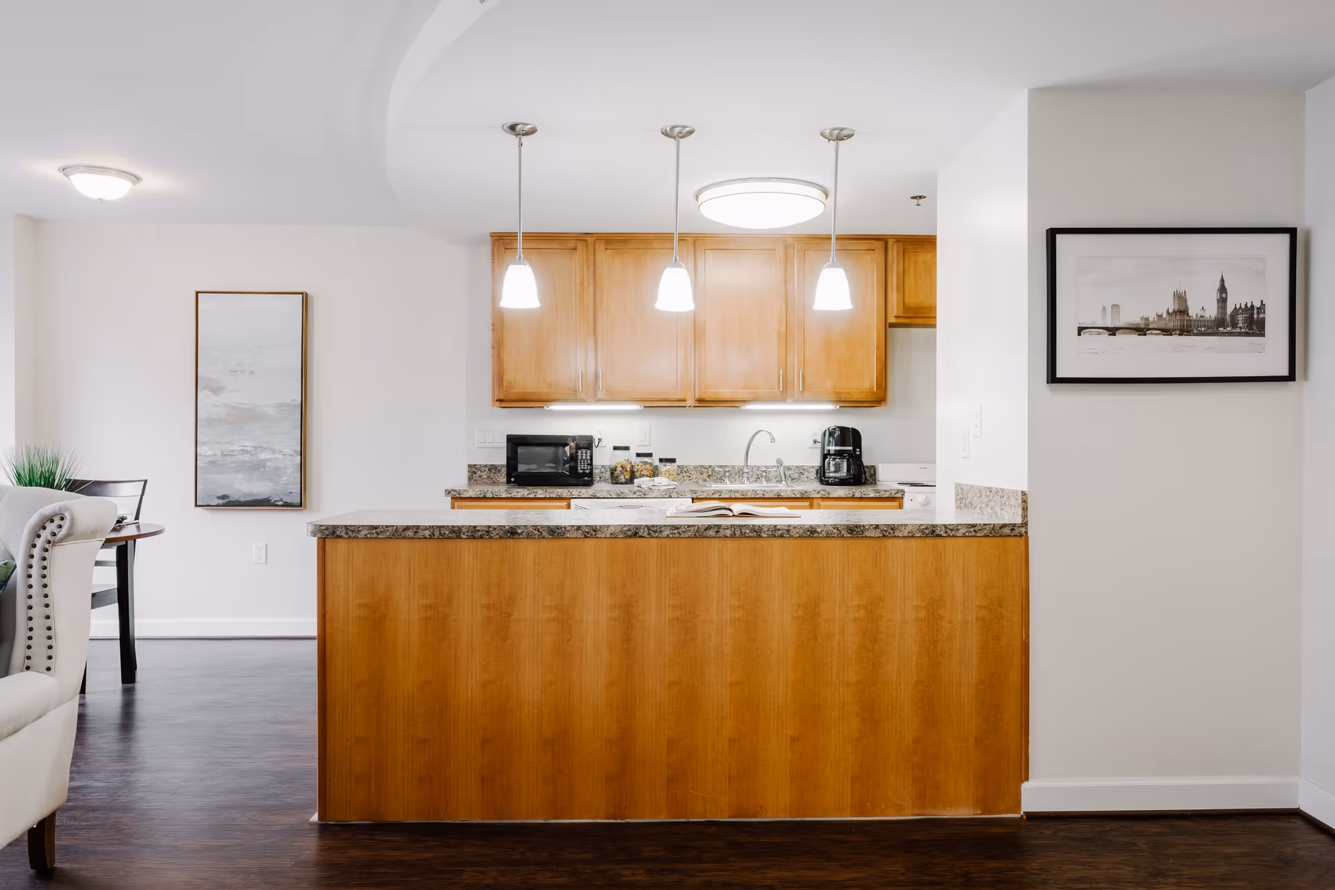 A modern kitchen area with wooden cabinets and a granite countertop island. Three pendant lights hang above the island. On the countertop, there is a microwave, an air fryer, and some jars. The kitchen is part of an open floor plan with a glimpse of a dining area and a white armchair on the left. A framed black and white picture hangs on the wall to the right.