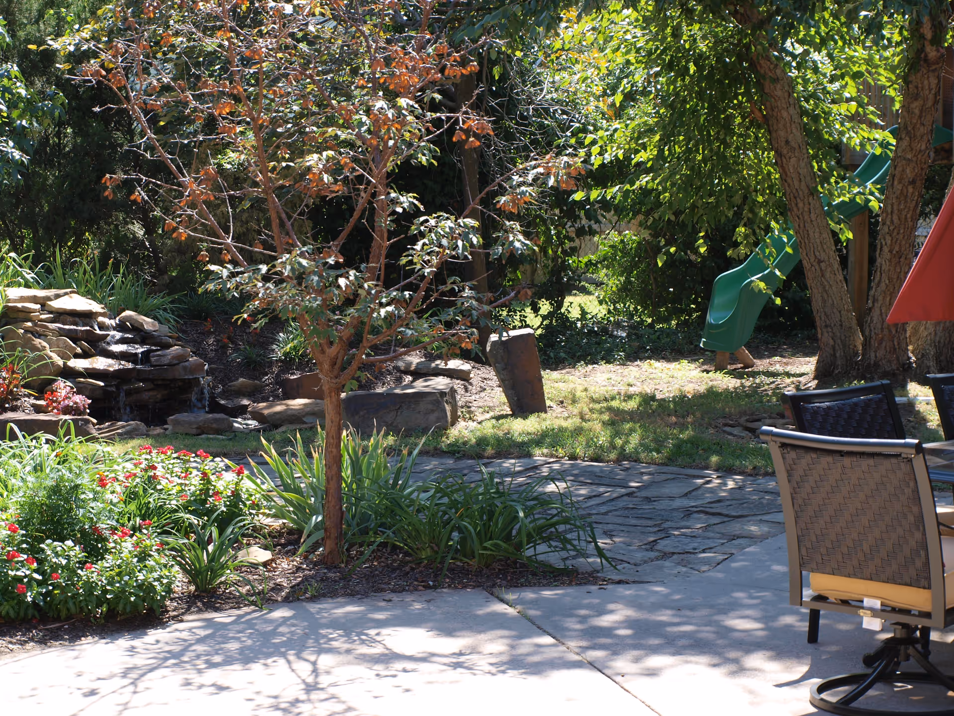Sunny courtyard with a small tree, flowering beds, a stone water feature, patio chairs, and a green slide.