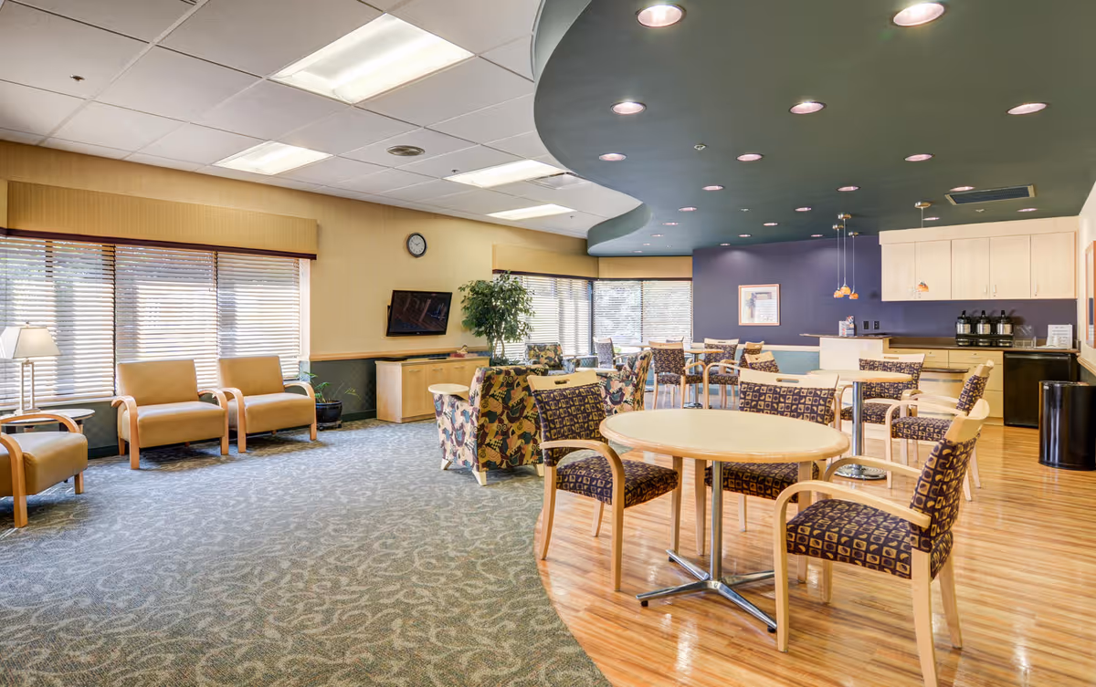 A spacious and well-lit common area in a healthcare and rehabilitation center featuring a mix of seating options including patterned armchairs around round tables, plain armchairs along the window, and floral armchairs near a large window. The room has a combination of carpet and wood flooring, a curved ceiling design with recessed lighting, a wall-mounted TV, a clock, and a small kitchenette area with cabinets, coffee machines, and a trash bin.