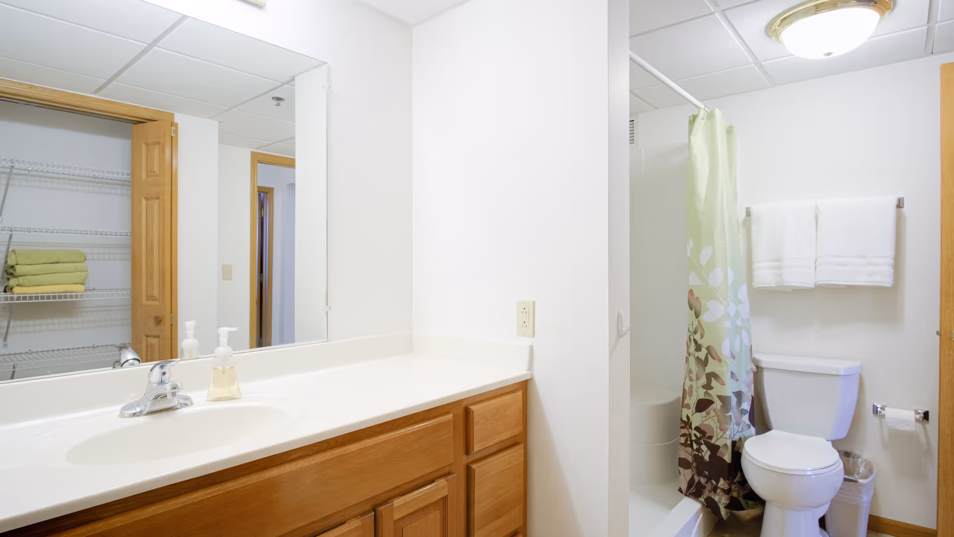 Bright bathroom with a wooden vanity and sink, large mirror, toilet, shower with a patterned curtain, and a towel rack.